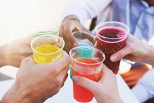 Detail on the hands of a group of young people holding colorful carbonated fruit drinks in plastic cups, with summer sunlight filtering through.